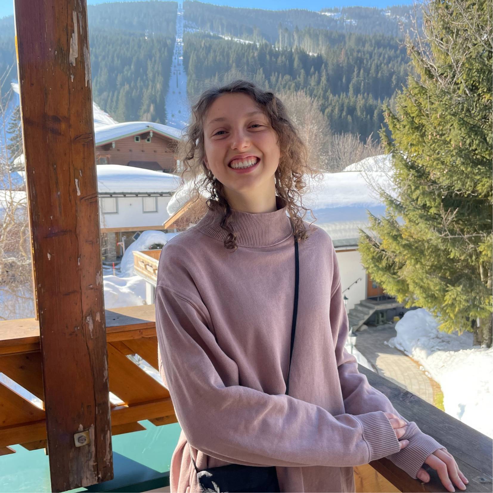 Image of a female smiling at the camera posing in front of a window overlooking a snow covered building and tree lined hill.
